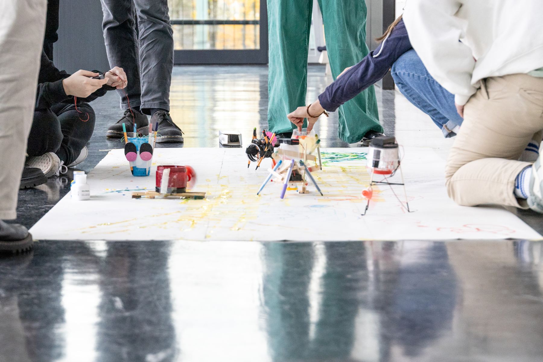 Robots drawing on a long sheet of paper on the floor, students kneeling around