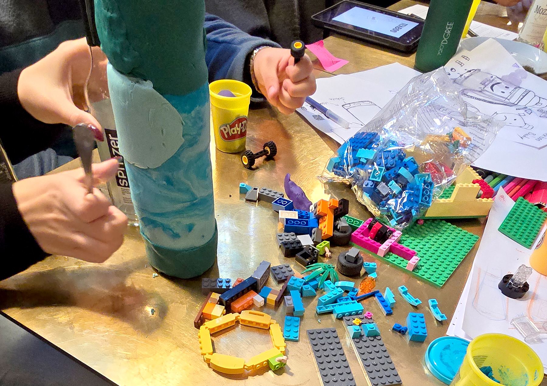 Hands shaping teal modelling clay around the bottle, LEGO bricks on the table